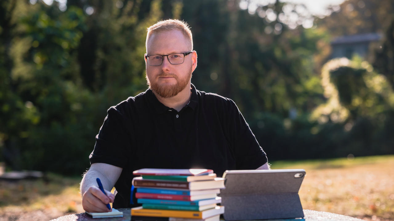 Foto von Sebastian Pertsch. Er sitzt arbeitend in einem Berliner Park und schaut konzentriert in die Kamera. Vor ihm ein Stapel Bücher, ein Tablet und ein Notizbuch, in das er scheinbar etwas schreibt. Er hat rote, kurze Haare, die zu einem Seitenscheitel gestylt sind, und er trägt einen Vollbart, eine eckige, leicht abgerundete Brille und ein schwarzes Poloshirt.