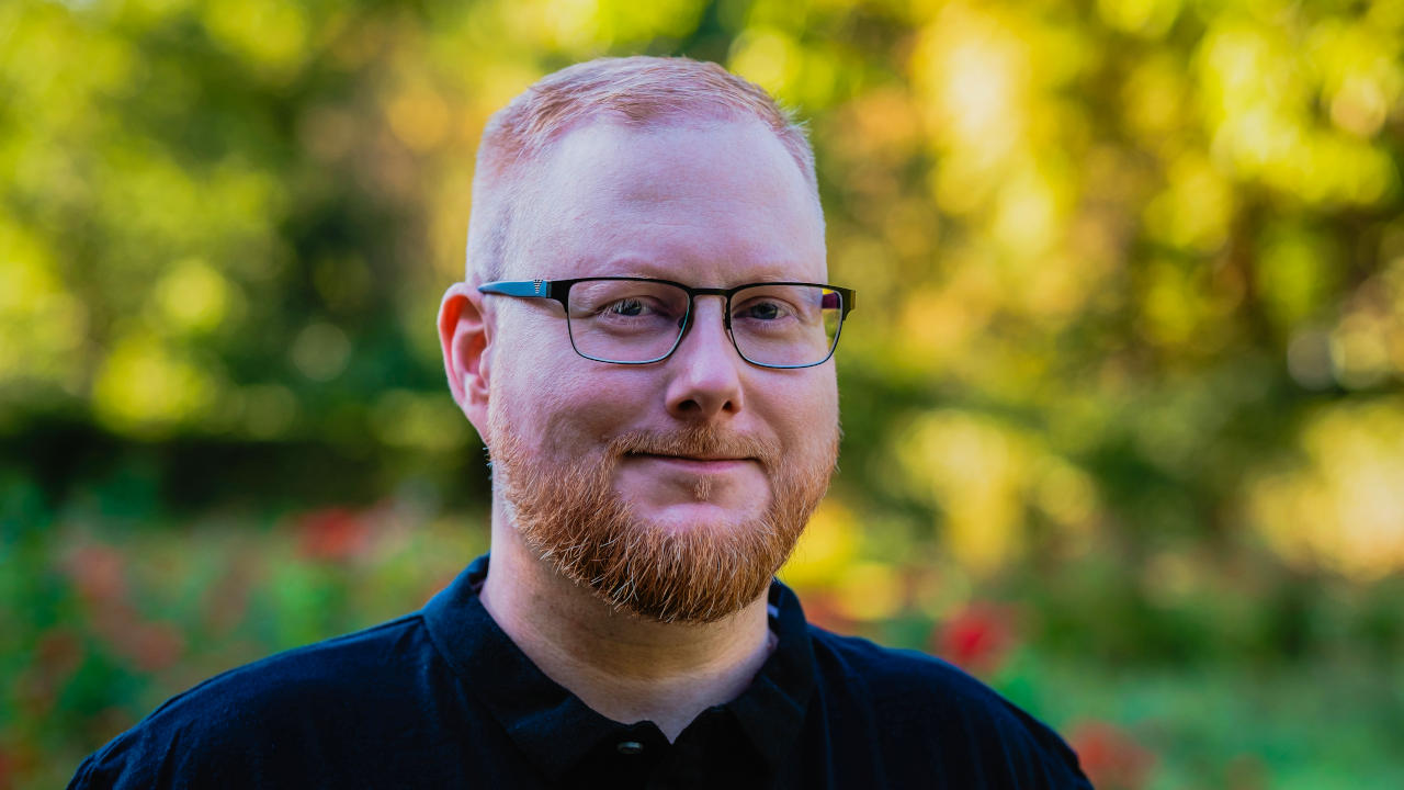 Foto von Sebastian Pertsch. Er steht vor einem Rosengarten und lächelt mit geschlossenem Mund in die Kamera. Er hat rote, kurze Haare, die zu einem Seitenscheitel gestylt sind, und er trägt einen Vollbart, eine eckige, leicht abgerundete Brille und ein schwarzes Poloshirt.