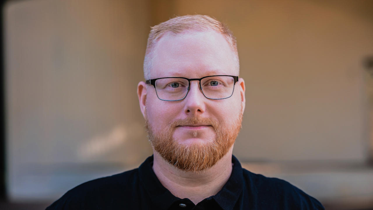 Foto von Sebastian Pertsch. Er schaut freundlich in die Kamera. Er hat rote, kurze Haare, die zu einem Seitenscheitel gestylt sind, und er trägt einen Vollbart, eine eckige, leicht abgerundete Brille und ein schwarzes Poloshirt.
