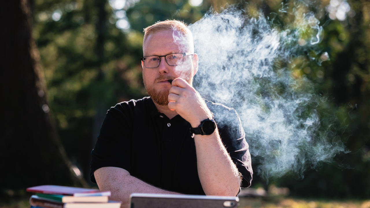 Foto von Sebastian Pertsch. Er sitzt in einem Berliner Park und raucht Tabakpfeife. Dabei schaut er konzentriert und nachdenkend in die Kamera. Er hat rote, kurze Haare, die zu einem Seitenscheitel gestylt sind, und er trägt einen Vollbart, eine eckige, leicht abgerundete Brille und ein schwarzes Poloshirt.