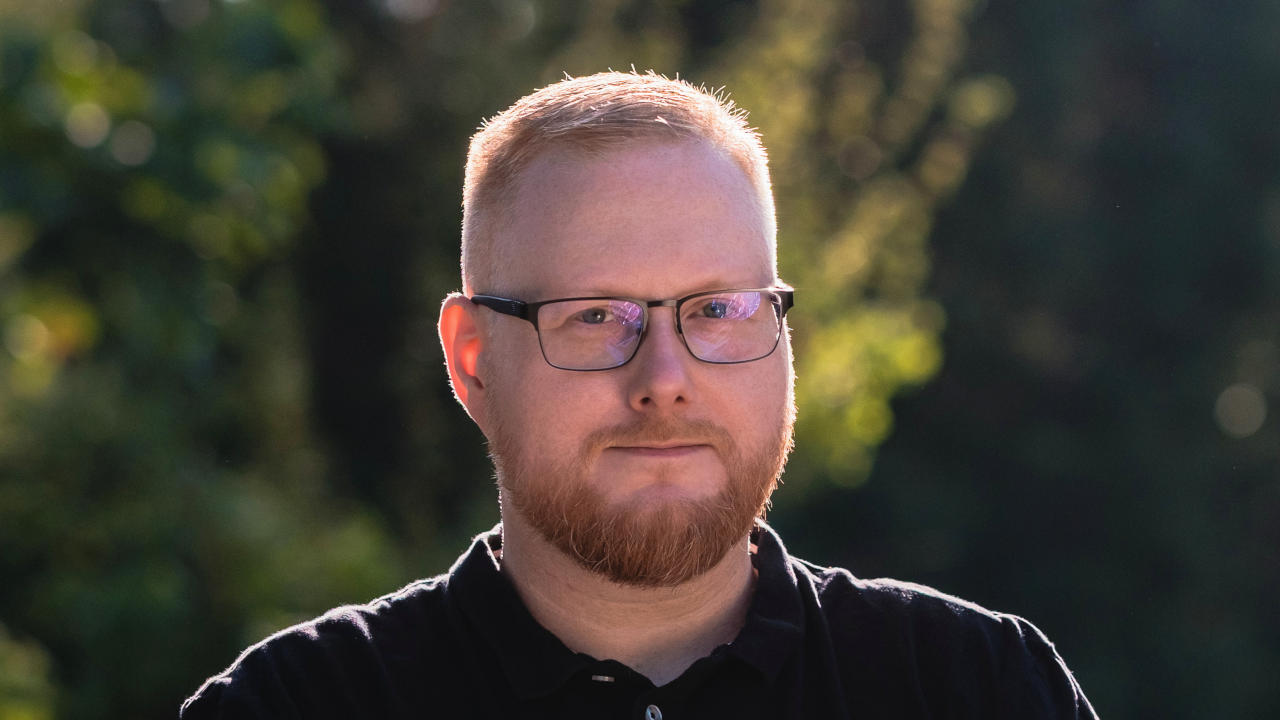 Foto von Sebastian Pertsch. Er steht vor Bäumen und schaut neutral in die Kamera. Die Sonne scheint von hinten und umgibt ihn mit einem hellen Schein. Er hat rote, kurze Haare, die zu einem Seitenscheitel gestylt sind, und er trägt einen Vollbart, eine eckige, leicht abgerundete Brille und ein schwarzes Poloshirt.